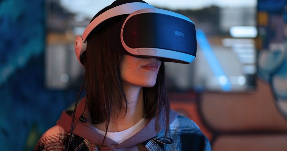 A girl wearing a virtual reality headset indoors. The headset is black with white accents and appears to be equipped with headphones for an immersive experience. The individual is indoors, as indicated by the soft lighting and furniture in the background.