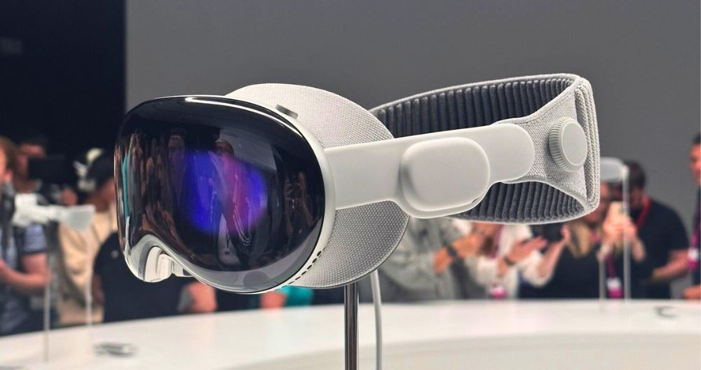 This is an image of the Apple Vision Pro virtual reality headset. The headset is predominantly white with a reflective, dark visor. It is mounted on a display stand against a blurred background. The design features textured fabric and an adjustable strap for comfort. Several people are in the background, seemingly interested in the device; some are taking photos.