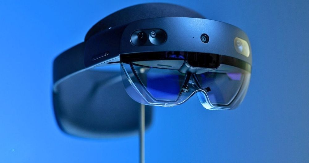 This is a Microsoft HoloLens 2, an augmented reality headset. It has a sleek design with dark visors and multiple sensors for an immersive experience. The image shows the device against a blue background.