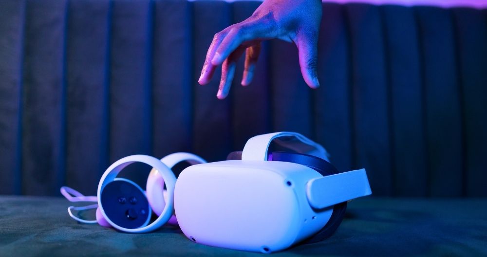 A hand reaching out towards a white virtual reality (VR) headset and headphones. The VR equipment is placed on a surface with a dark, textured backdrop that appears to be a cushioned seat or wall.