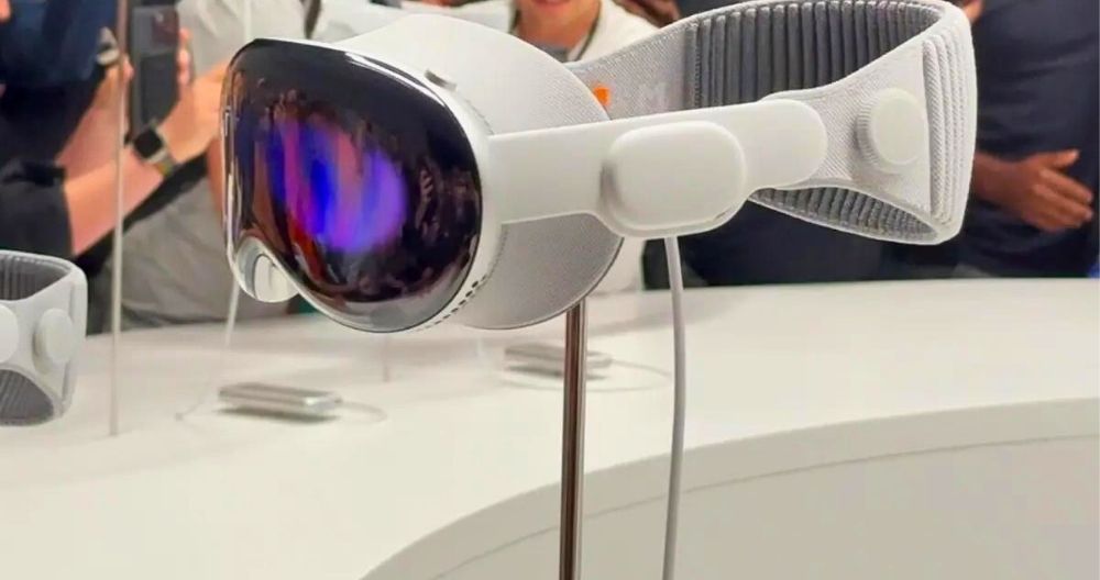 Apple Vision Pro is presented in exhibition by Apple. The headset is placed on a stand. Some people are visible in the background. 